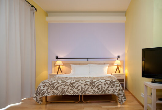 A hotel bedroom, modern furniture and fittings, double bed and bedside lights, television and long white curtains.
