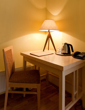 A room with a small desk and chair with a lit table lamp, tray and kettle and telephone. 