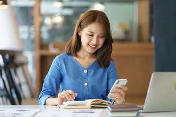 Charming young Asian businesswoman with a smile sitting holding smartphone at the office.