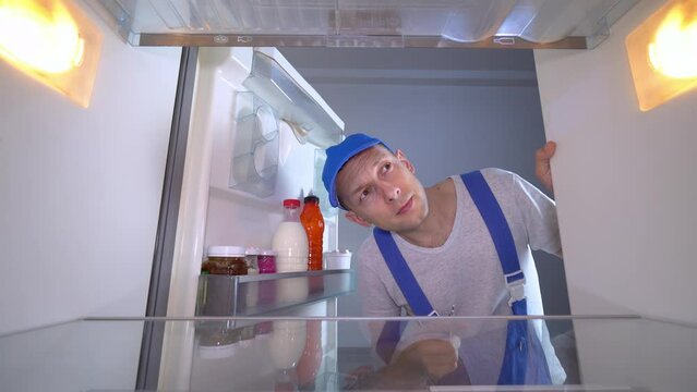 The refrigerator repairman opens the door and examines the inside of the refrigerator with a screwdriver in his hand. A young Caucasian man in a blue jumpsuit or uniform. Refrigerator repair.