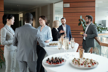 Two groups of successful speakers in formalwear talking by served tables during buffet in luxurious...