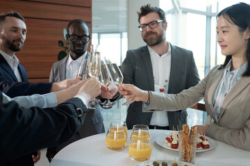 Happy interracial entrepreneurs clinking with flutes of champagne over table served with glasses of juice, canape and other snacks - obrazy, fototapety, plakaty