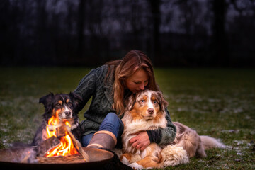 Young woman is sitting outside in the woods with her two Australian Shepherd dogs. Snow on the grass, at night by the campfire. She kisses the dog lovingly