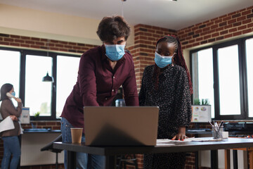 Multi ethnic diverse office workers wearing facemasks against coronavirus analyzing company accounting data and reviewing partner portfolio. Expectant mother finance advisor looking over project ideas