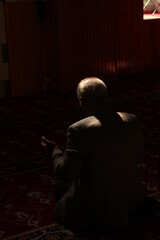 Obraz na płótnie Canvas Back of unrecognizable white old man praying in dark mosque with a beam of sunlight falling over head and shoulders. Prayer area with red rug, carpet and vintage lights.