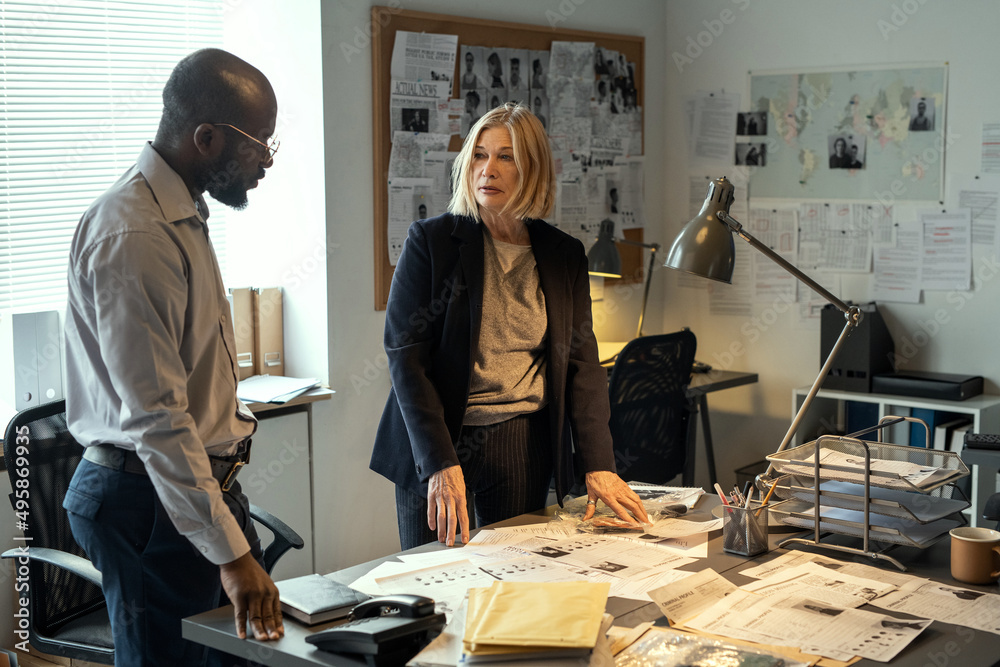 Poster two interracial fbi agents standing by desk with profiles of criminals and discussing their ideas ab