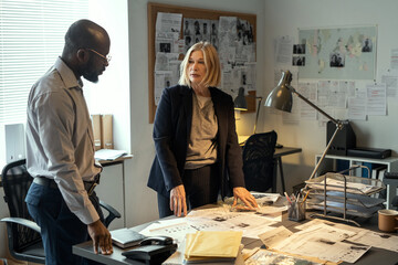 Two interracial FBI agents standing by desk with profiles of criminals and discussing their ideas...
