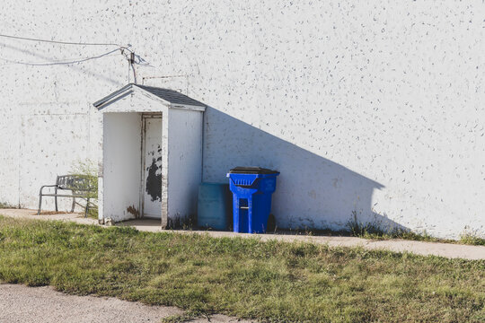 The white wall of a building in a small town, blue refuse bin and bench. 