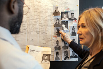 Blond confident female detective pointing at photo of suspect on board over map while matching facts of serial crimes with colleague - obrazy, fototapety, plakaty