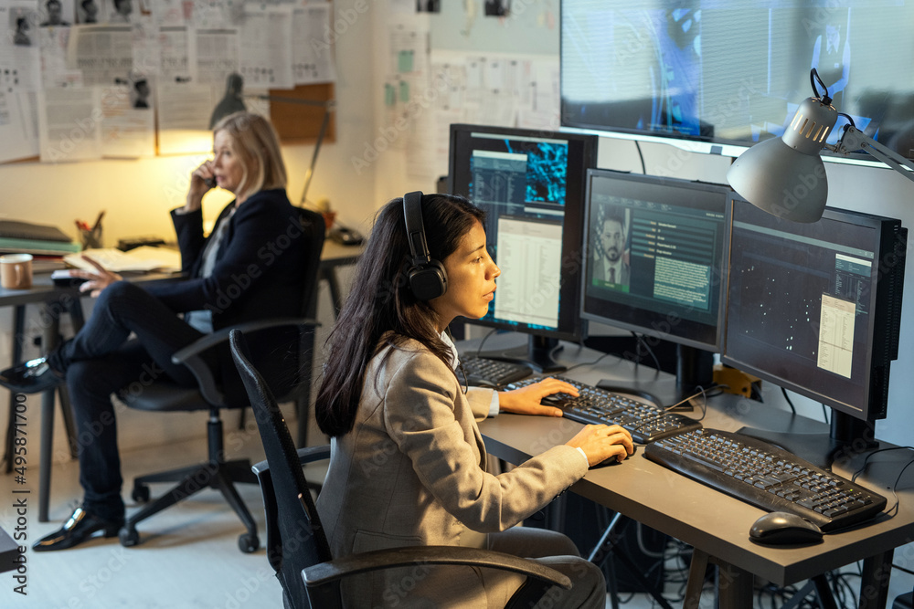 Poster serious female agent of intelligence service sitting in front of computer monitors in office and pro
