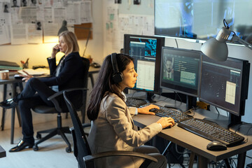 Serious female agent of intelligence service sitting in front of computer monitors in office and...