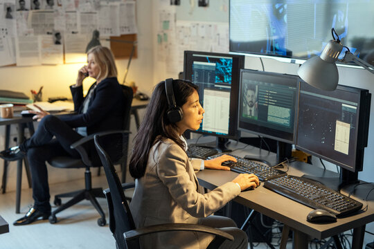 Serious female agent of intelligence service sitting in front of computer monitors in office and processing personal data of criminals