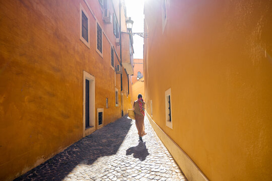 Fototapeta Narrow old street with orange buildings and woman walking against the sun. Woman traveling in Rome, Italy. Wide angle view