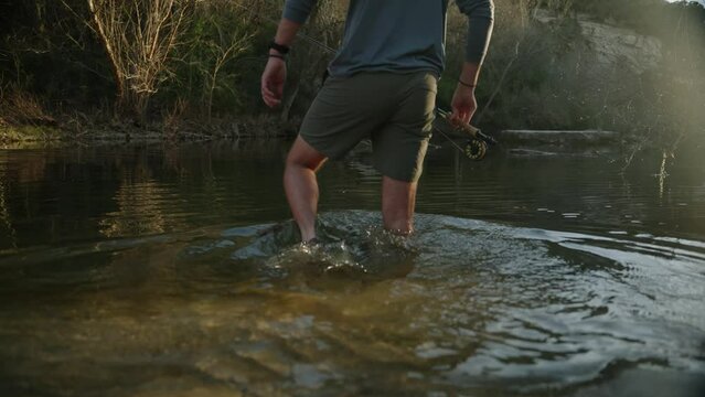 Male fly fisherman steps into sun soaked lake water with sandals close up slow motion