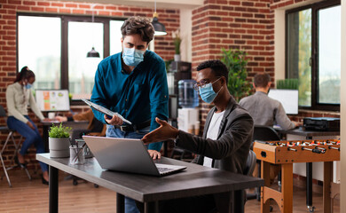 Fototapeta na wymiar Diverse businessmen working at marketing project analyzing company graph discussing business ideas in brick wall startup office. Multi-ethnic team wearing medical face mask against covid19