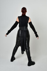 Full length portrait of pretty red haired female model wearing black futuristic scifi leather costume. Dynamic standing poses with gestural hands, facing backwards away from  on a white studio backgro