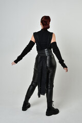 Full length portrait of pretty red haired female model wearing black futuristic scifi leather costume. Dynamic standing poses with gestural hands, facing backwards away from  on a white studio backgro