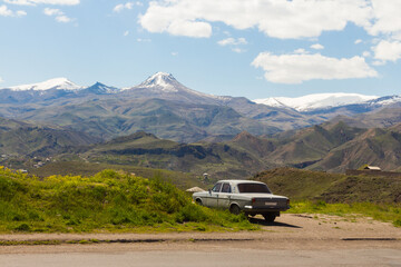 An old Soviet car stands on the roadside against the backdrop of snow-capped mountain peaks. Armenia