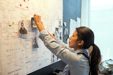 Young Hispanic female agent of FBI in uniform pinning photos of several male suspects on map while suggesting location of their gang - obrazy, fototapety, plakaty