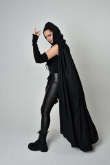 Naklejka premium Full length portrait of pretty red haired female model wearing black futuristic scifi leather costume with black flowing cloaked cape. Dynamic standing poses with gestural hands, facing backwards away
