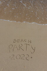 Fototapeta premium The inscription beach party 2022 on the sand by the water and the rising wave, seashore beach vacation by the sea
