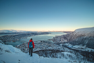 Female Adventurer in a red jacket and backpack stands atop a fjellheisen and watches Norwegian Sea surrounding the city of Tromso in northern Norway above the Arctic Circle on frosty winter morning