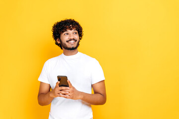 Positive indian or arabian guy, in white t-shirt, holding smartphone in hands, texting online, browsing internet, looking happily to the side, thinking, standing on isolated orange background, smiling