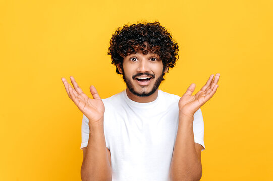 Surprised confused indian or arabian guy with curly hair, wearing white t-shirt, looking at camera in disbelief, spreading his arms to the sides, shrugging, standing over isolated orange background