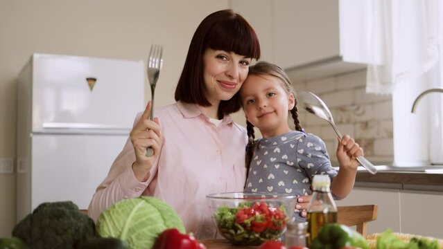 Young mother hugging her child daughter and looking at each other holding a spoon and fork while preparing proper meal with vegetables. Happy girl assisting mother in preparing salad