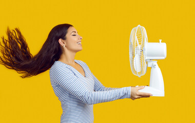 Profile view of happy young woman standing isolated on yellow background, holding electric fan,...