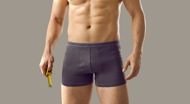 Does size matter. Man with fit, attractive body and naked torso measuring his big penis with centimeter tape. Sexy man in boxer briefs holding yellow metal meter standing isolated on grey background
