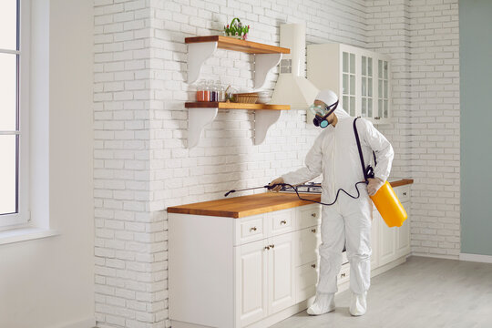 Exterminator fighting termites or cockroaches in the house. Pest control guy in mask and protective white suit spraying insecticide from sprayer bottle on cupboards and countertop in kitchen interior