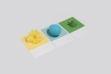 Minimal spring concept with blue, yellow and green cards, Easter egg, daisy flower and plant leaf. Trendy colorful April concept.