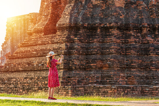 solo traveler concept. happy Asian woman tourist in red dress taking picture of old pagoda around history temple for cultural tourism at Ayutthaya World Heritage Site, Thailand