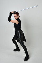 Full length portrait of pretty red haired female model wearing black futuristic scifi leather costume, holding a lightsaber sword weapon. Dynamic standing poses  on a white studio background.