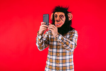 Man with chimpanzee monkey mask and plaid shirt, consulting his mobile phone, on red background.