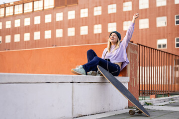 young blonde woman sitting on a wall listening music, female skater having relax in the city, happy...