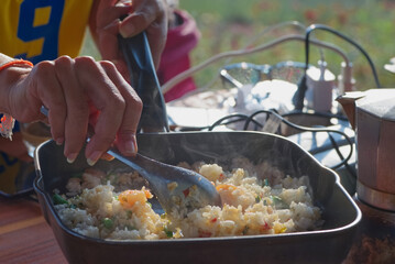 A person is cooking fried rice in a pan with blur green field background at a morning