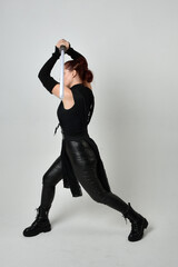 Obraz na płótnie Canvas Full length portrait of pretty red haired female model wearing black futuristic scifi leather costume, holding a lightsaber sword weapon. Dynamic standing poses on a white studio background.