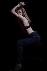Fototapeta na wymiar Sexy and beautiful young blonde with a slender body, long seductive legs posing in jeans on a chair, dark wall on the background