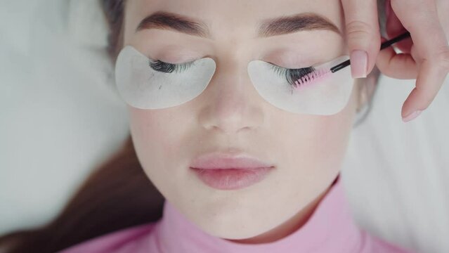 Lashmaker combs the eyelashes of a beautiful young woman with a brush