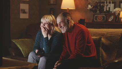 Happy senior husband and wife laughing at funny joke while sitting on couch and watching comedy on TV at night at home