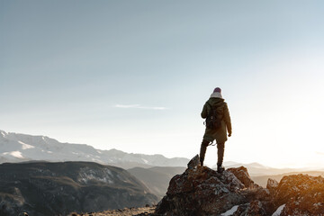 Young girl hiker stands on big rock against the backdrop of mountains and watches the sunset