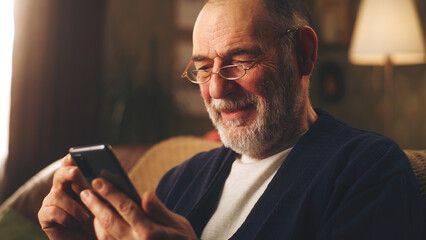 Elderly man smiling and watching funny videos on social networks using smartphone and resting in the evening sitting on the couch