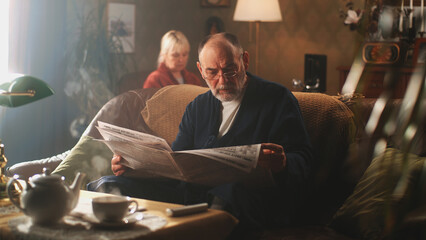 Aged man in bathrobe and glasses sitting on couch and reading daily newspaper in cozy living room, near woman in morning at home