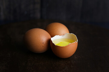 Fresh brown eggs and a broken egg with yolk on a dark wooden background