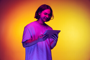 Portrait of young man with long hair using digital gadget isolated on orange background in purple...