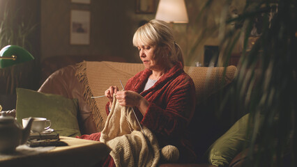 A retired woman knitting a warm sweater at her leisure time while sitting on the sofa in the...