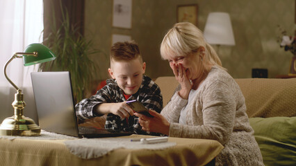 Teenager with elderly grandmother playing smartphone and laptop together at home sitting on the...