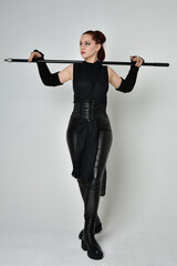 Full length portrait of pretty red haired female model wearing black futuristic scifi leather costume, holding a staff spear weapon. Dynamic standing pose on a white studio background.